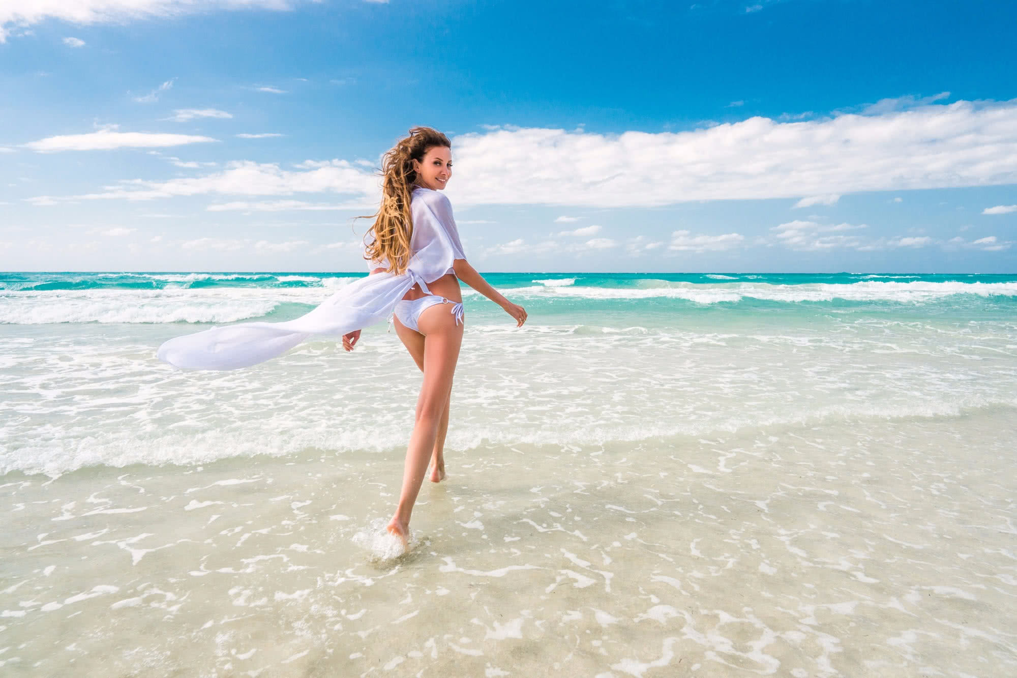 Beautiful young woman with long blond hair in white dress and swimsuit is running on the sand of caribbean coastline at summer sunset